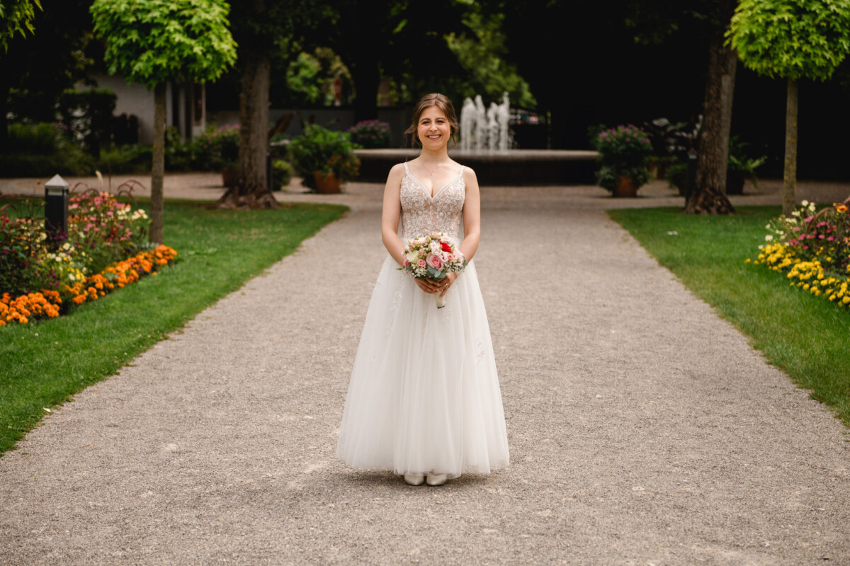 Eine Frau in einem weißen Hochzeitskleid hält einen Blumenstrauß und steht auf einem Kiesweg in einem Garten mit Bäumen und Blumen.