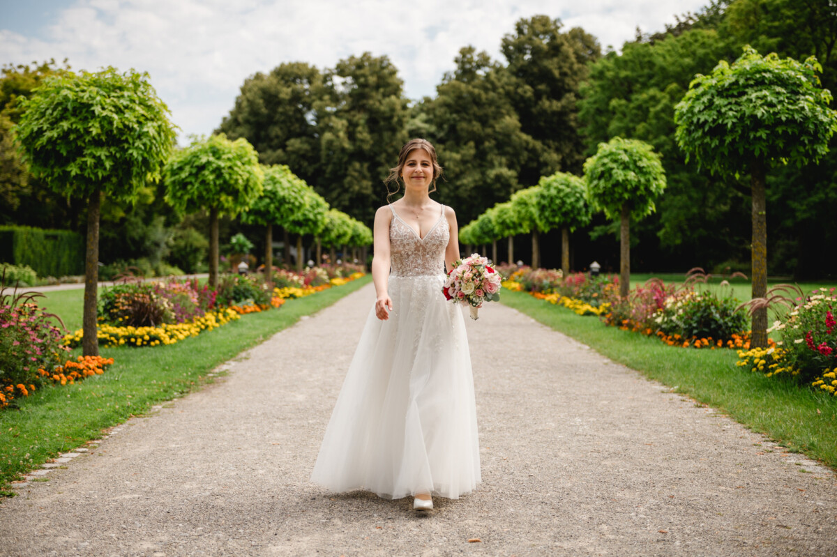Eine Braut in einem weißen Kleid hält einen Blumenstrauß, während sie einen blumengesäumten Weg in einem Garten entlanggeht.