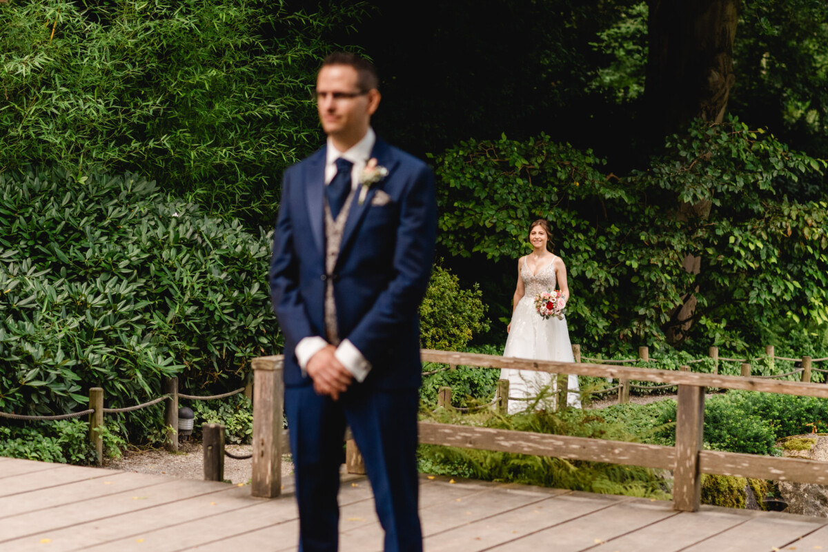 Eine Braut im weißen Kleid und mit einem Blumenstrauß in der Hand nähert sich einem Bräutigam im blauen Anzug, der auf einem Holzdeck steht, umgeben von üppigem Grün.