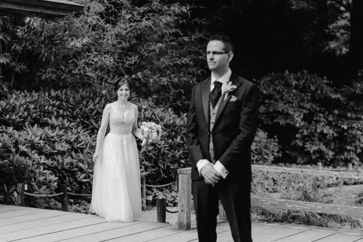 Auf einem Schwarzweißfoto nähert sich die Braut dem Bräutigam, der auf einer Holzplattform steht, umgeben von Bäumen und Büschen.