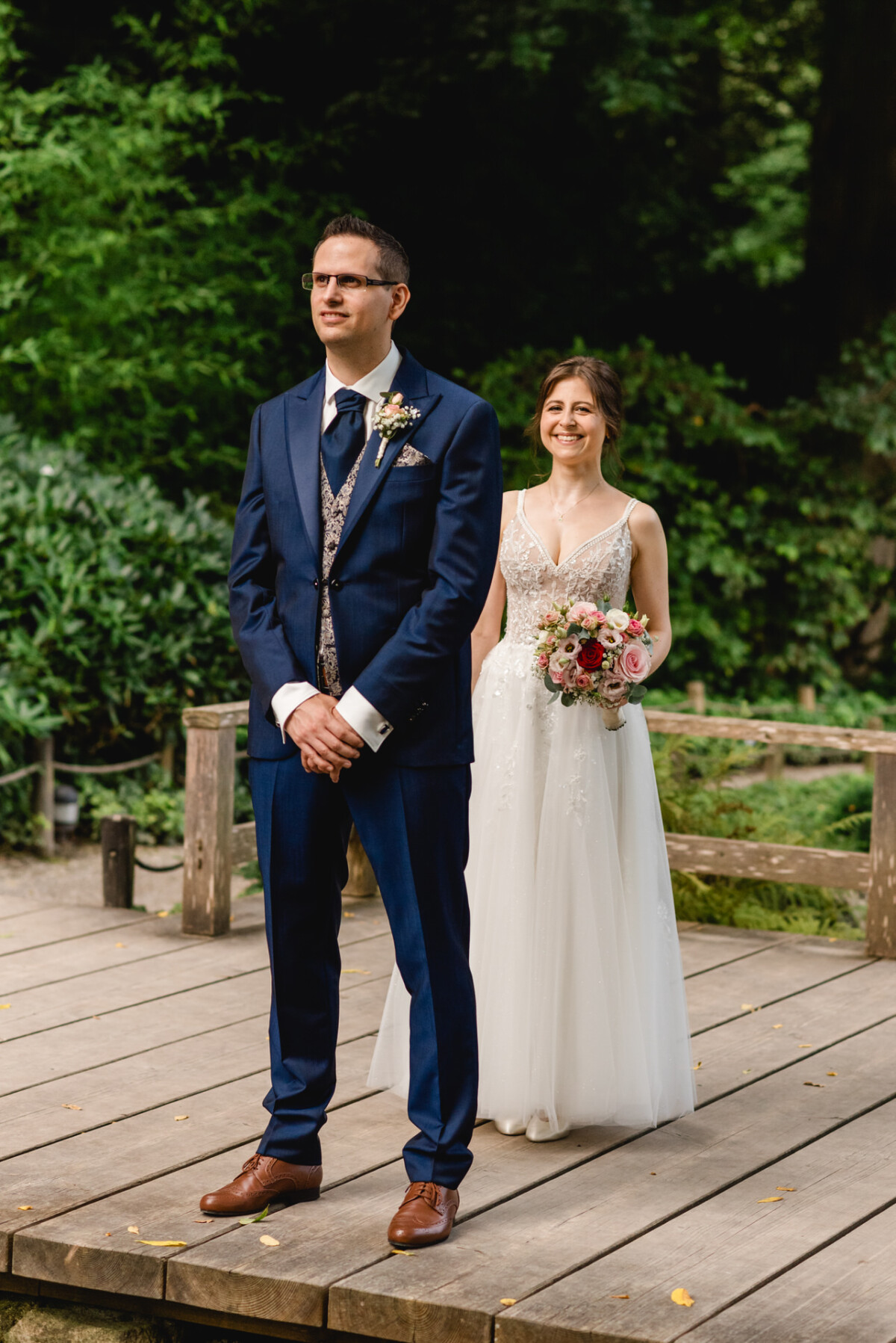 Braut und Bräutigam stehen auf einer Holzplattform im Freien. Der Bräutigam trägt einen blauen Anzug und die Braut hält einen Blumenstrauß und trägt ein weißes Kleid.