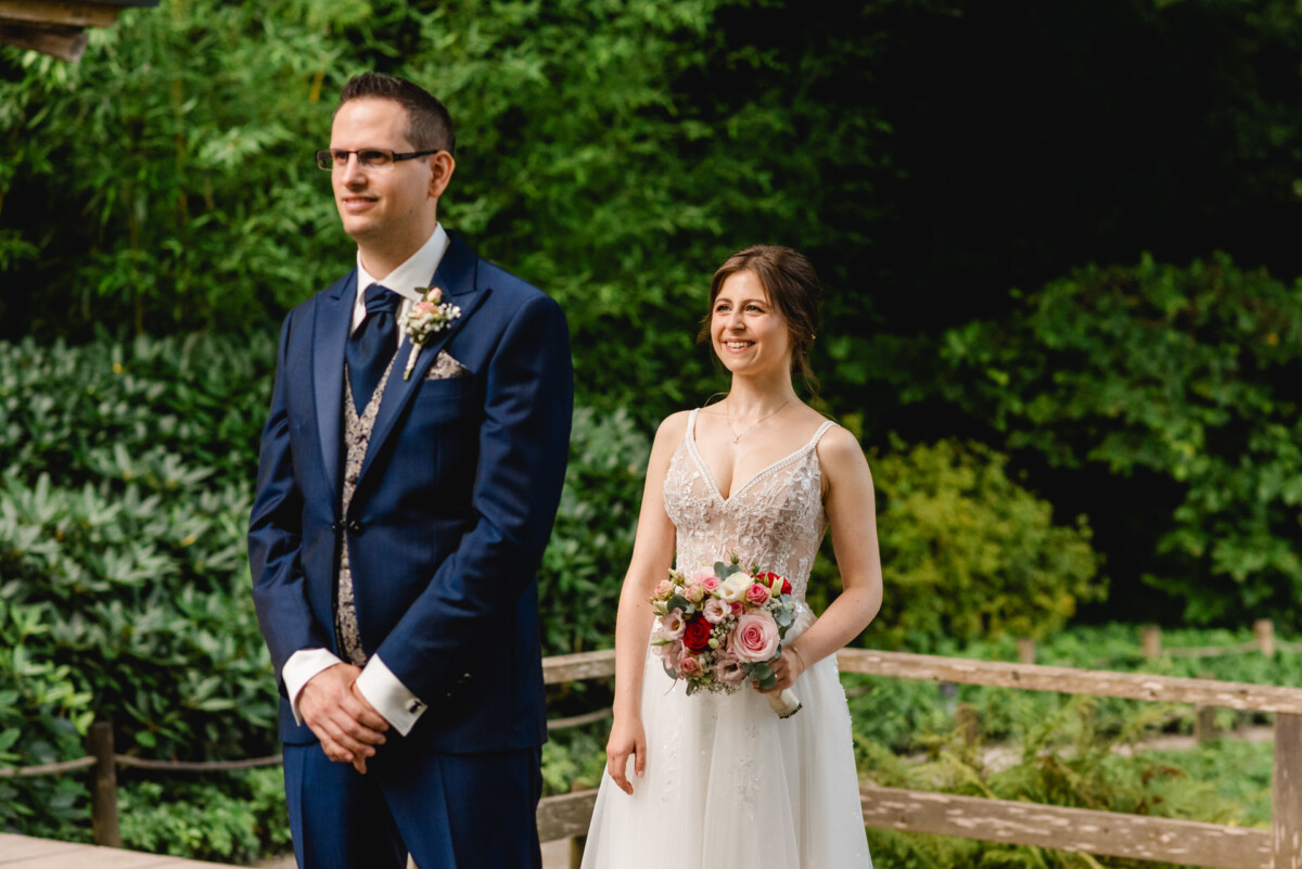 2024.07.27-Beatrice-Christian-Gut-Mergenthau-Kissing_00025 Eine Braut und ein Bräutigam stehen draußen. Die Braut hält einen Blumenstrauß und lächelt, während der Bräutigam geradeaus blickt. Sie sind von Grün umgeben.
