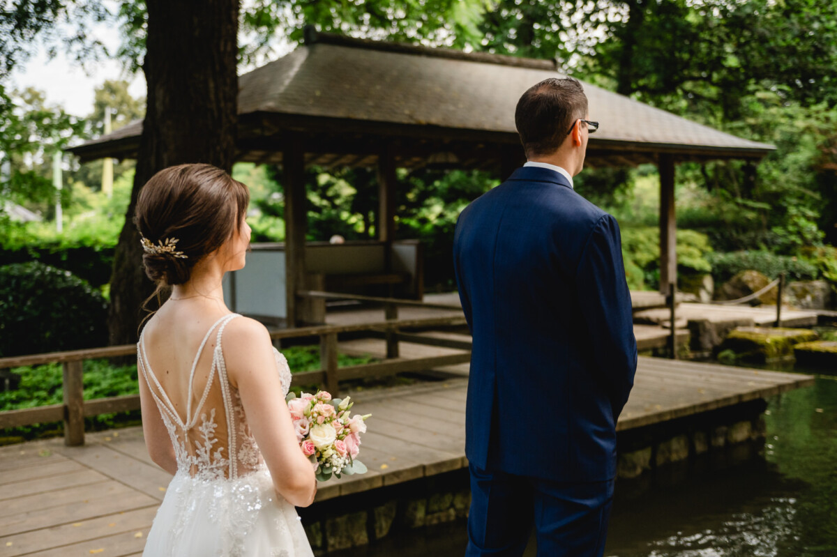 Braut und Bräutigam stehen auf einem Holzdeck neben einem Teich, wobei der Bräutigam abgewandt ist und die Braut einen Blumenstrauß hält.