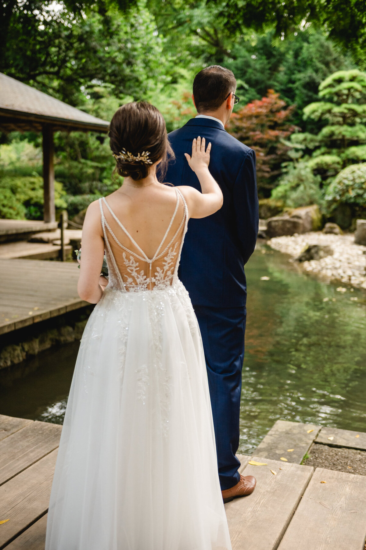 Eine Braut im weißen Kleid nähert sich einem Mann im marineblauen Anzug auf einer Holzbrücke in der Nähe eines Gartenteichs.