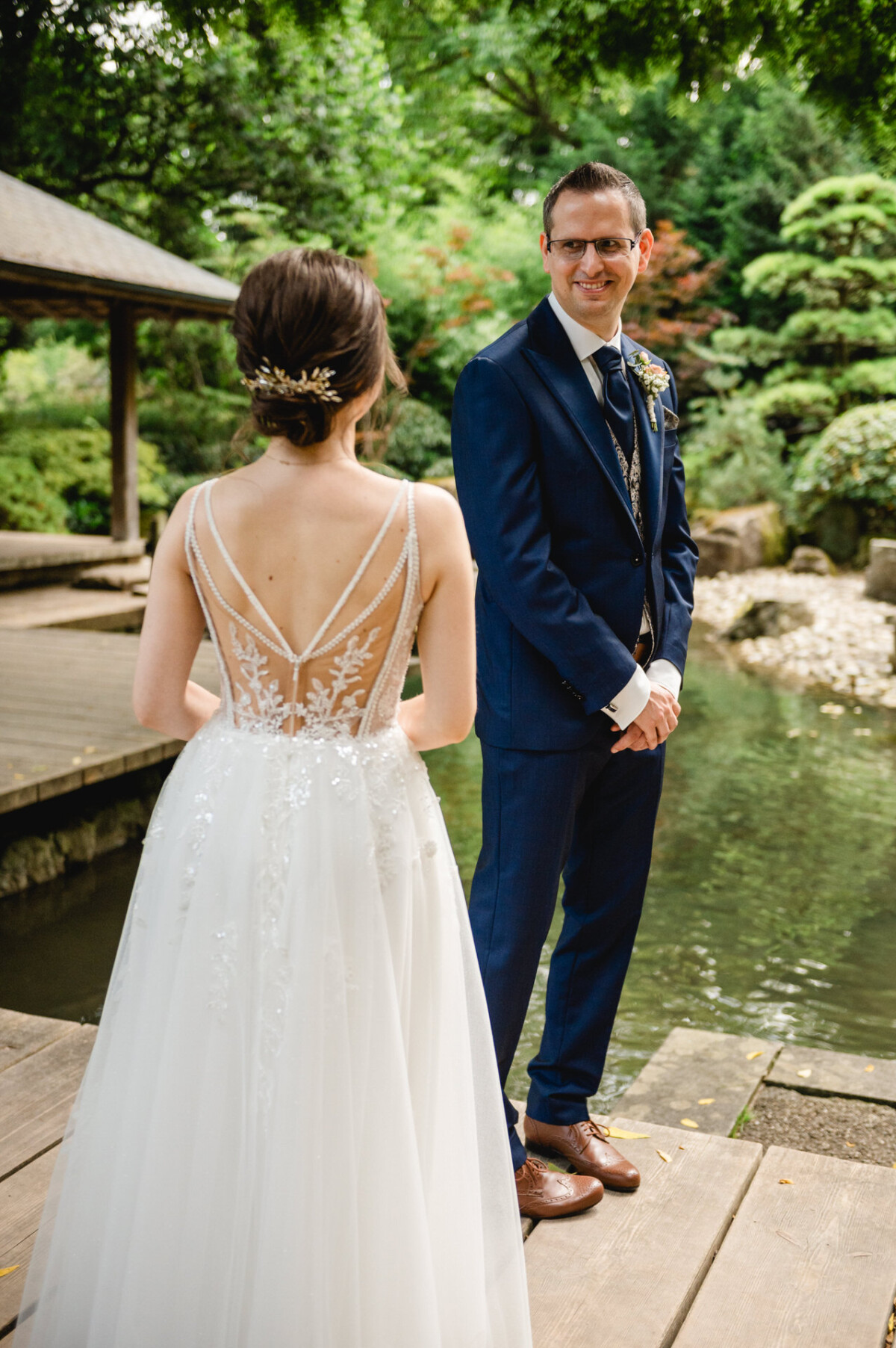 Ein Paar steht auf einem Holzweg neben einem Teich. Die Frau trägt ein weißes Kleid mit Spitzendetails und der Mann einen marineblauen Anzug. Sie sind von Bäumen und Grün umgeben.