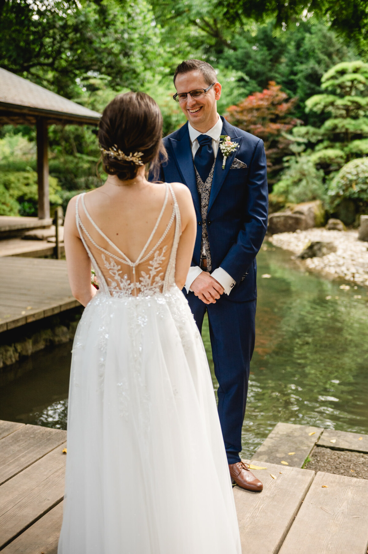 Ein Bräutigam im blauen Anzug lächelt eine Braut im Spitzenbrautkleid neben einem Teich in einem Garten an.