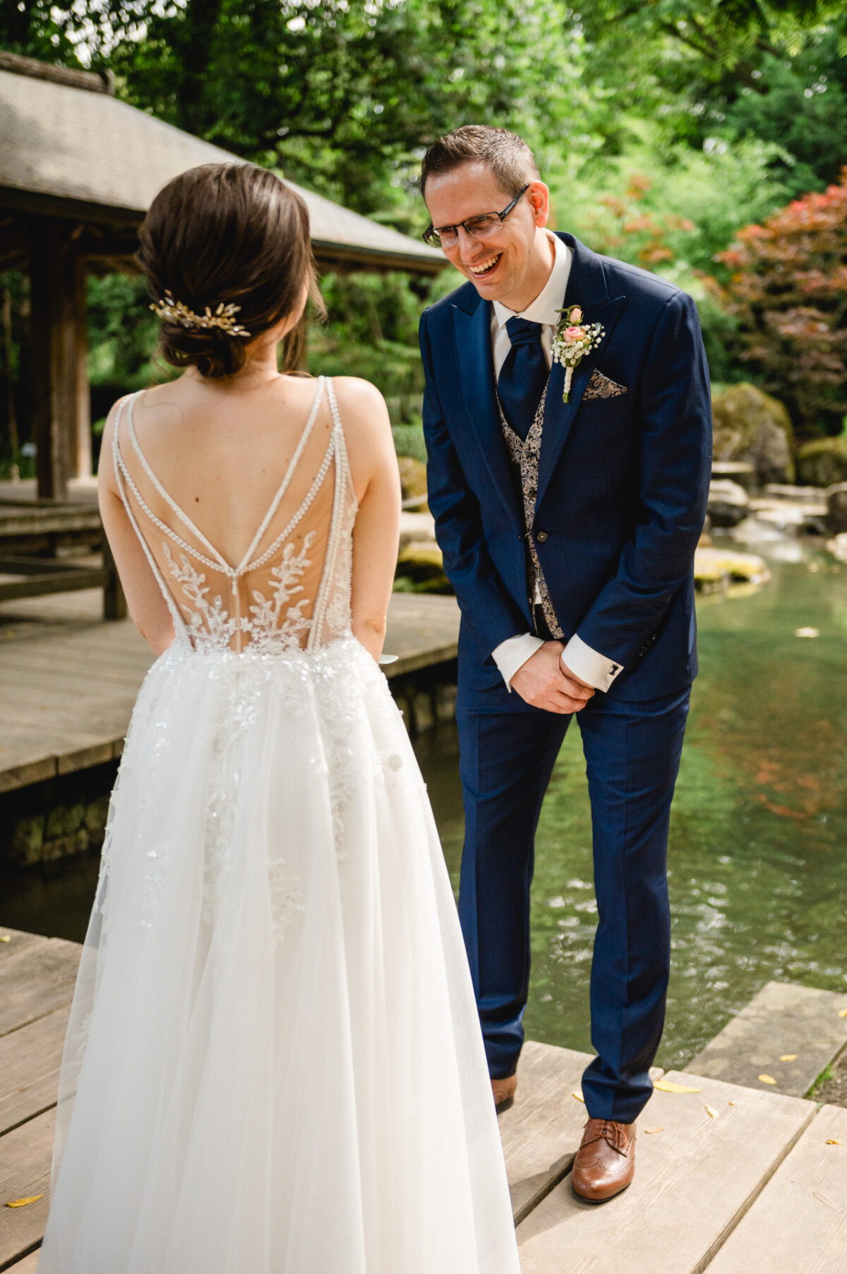 Braut und Bräutigam stehen auf einem Holzweg neben einem Teich. Der Bräutigam lächelt die Braut an, die ein weißes Kleid mit Spitzendetails und ein Haarteil trägt. Im Hintergrund sind Bäume und eine Holzkonstruktion zu sehen.
