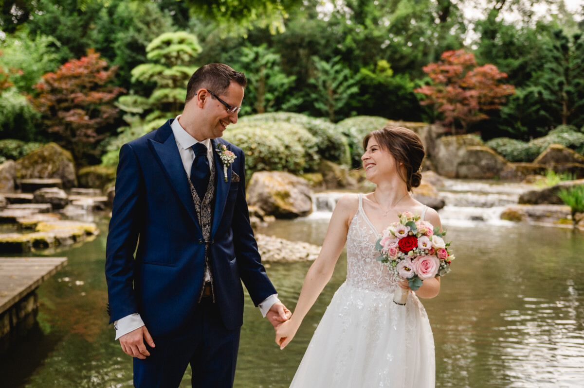 Braut und Bräutigam lächeln einander an, während sie vor einem Teich und Garten Händchen halten. Die Braut hält einen Strauß rosa und weißer Blumen.