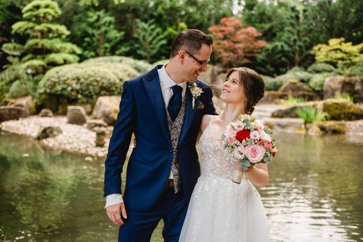 Braut und Bräutigam stehen an einem Teich in einem Garten. Die Braut hält einen bunten Blumenstrauß und sie lächeln sich an.