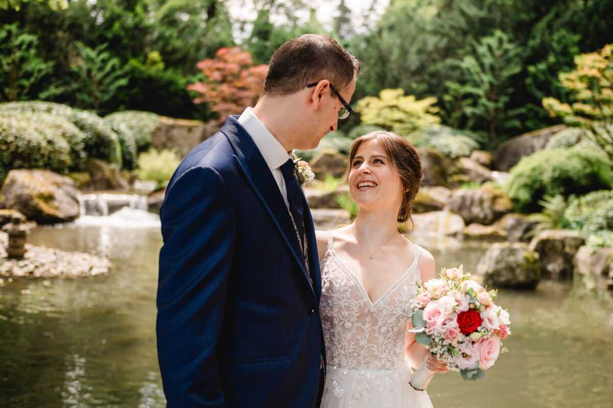 Ein Paar im Hochzeitskleid lächelt sich in einem Garten mit Teich und Grün im Hintergrund an. Die Braut hält einen Strauß rosa und roter Blumen.