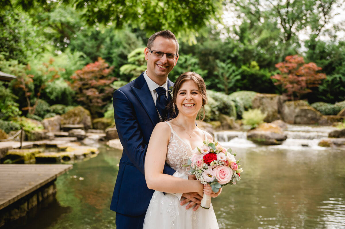 Ein Paar im Hochzeitskleid steht lächelnd vor einem Teich mit Grünflächen und Felsen im Hintergrund. Die Frau hält einen Blumenstrauß.