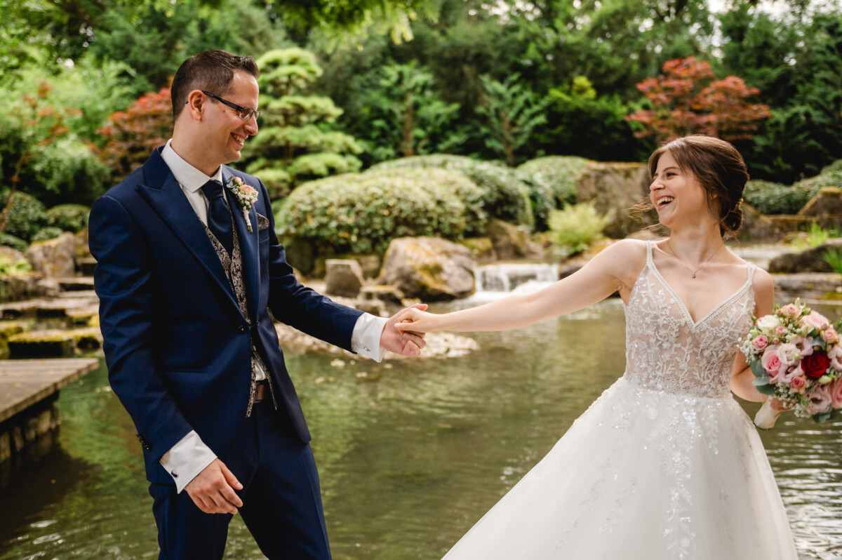 Eine Braut und ein Bräutigam halten Händchen und lächeln einander an, während sie an einem Teich in einem Garten stehen, wobei die Braut einen Strauß rosa und weißer Blumen hält.