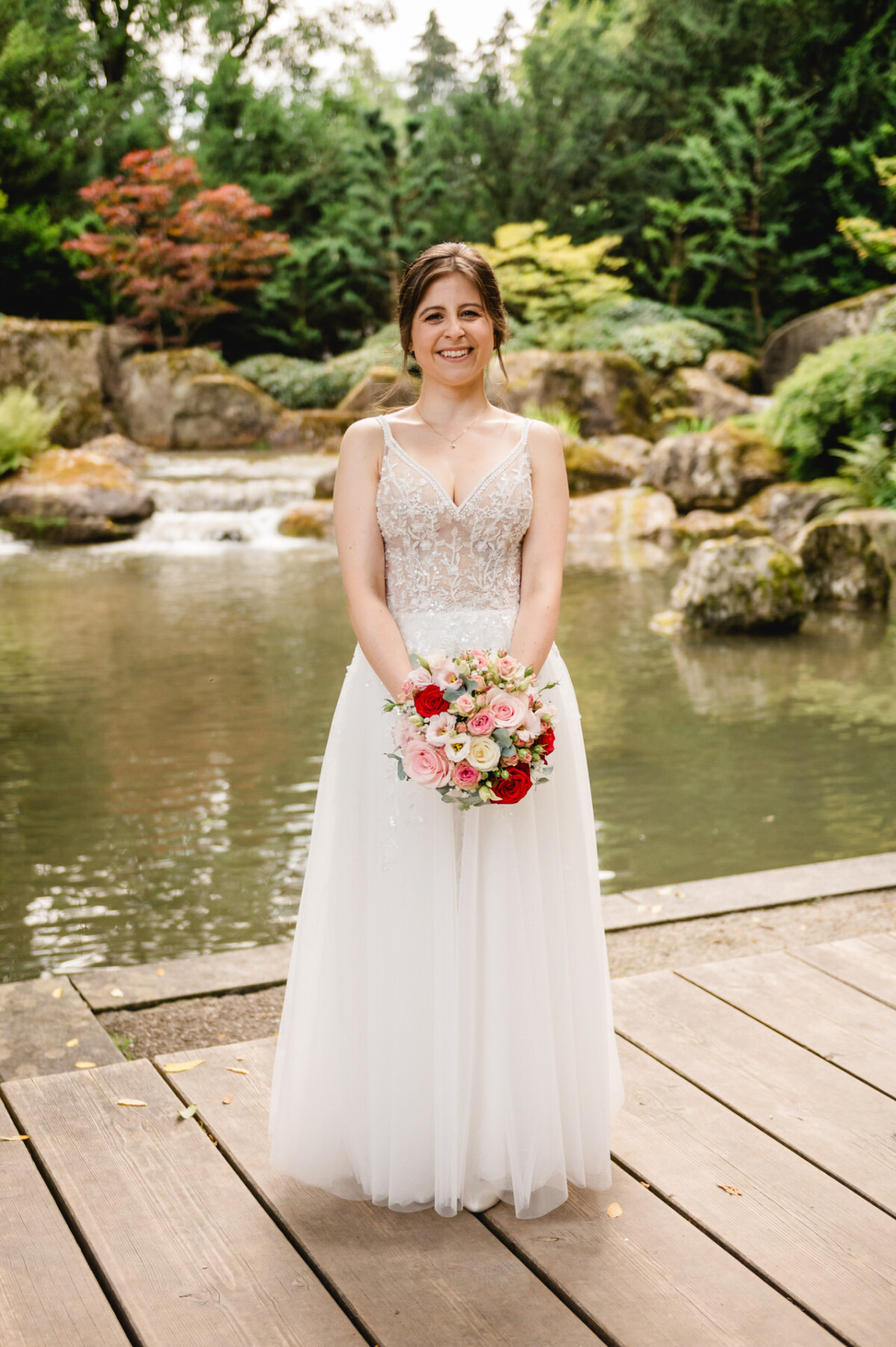 Braut in einem weißen Kleid hält einen Blumenstrauß und steht auf einem Holzweg neben einem Teich mit Felsen und Grün im Hintergrund.