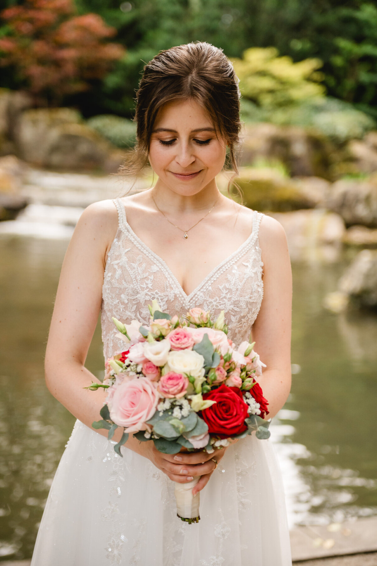 Eine Frau in einem weißen Spitzenkleid hält einen bunten Blumenstrauß, während sie vor einem ruhigen Teich und Garten steht.