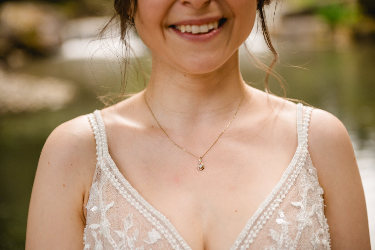 Eine Frau mit Halskette und Spitzenkleid lächelt im Freien am Wasser.