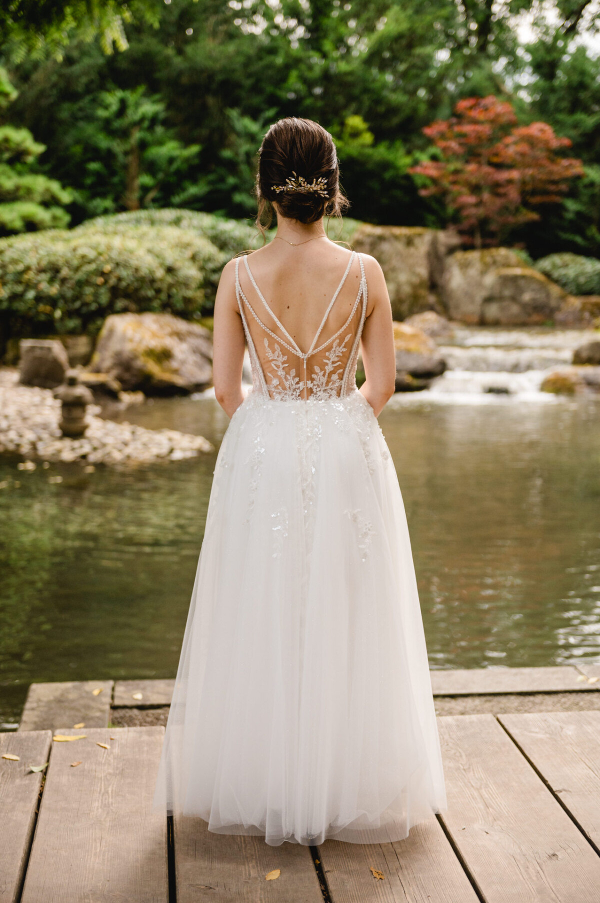 Eine Frau in einem weißen Kleid mit aufwendigen Rückendetails steht auf einer Holzterrasse mit Blick auf einen Teich und eine Gartenlandschaft.