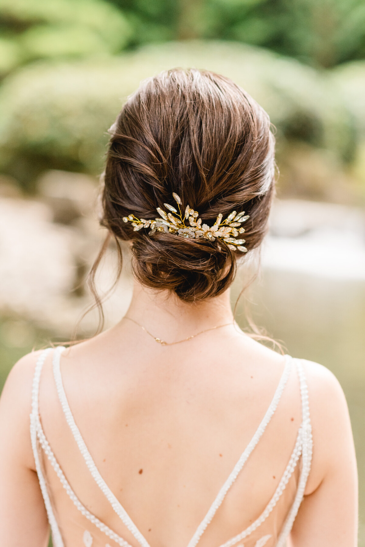 Nahaufnahme einer Frau mit brünettem Haar, das zu einem tiefen Dutt gestylt und mit einem dekorativen Haarteil geschmückt ist. Sie trägt ein weißes Kleid mit schmalen Trägern, im Freien mit viel Grün im Hintergrund.