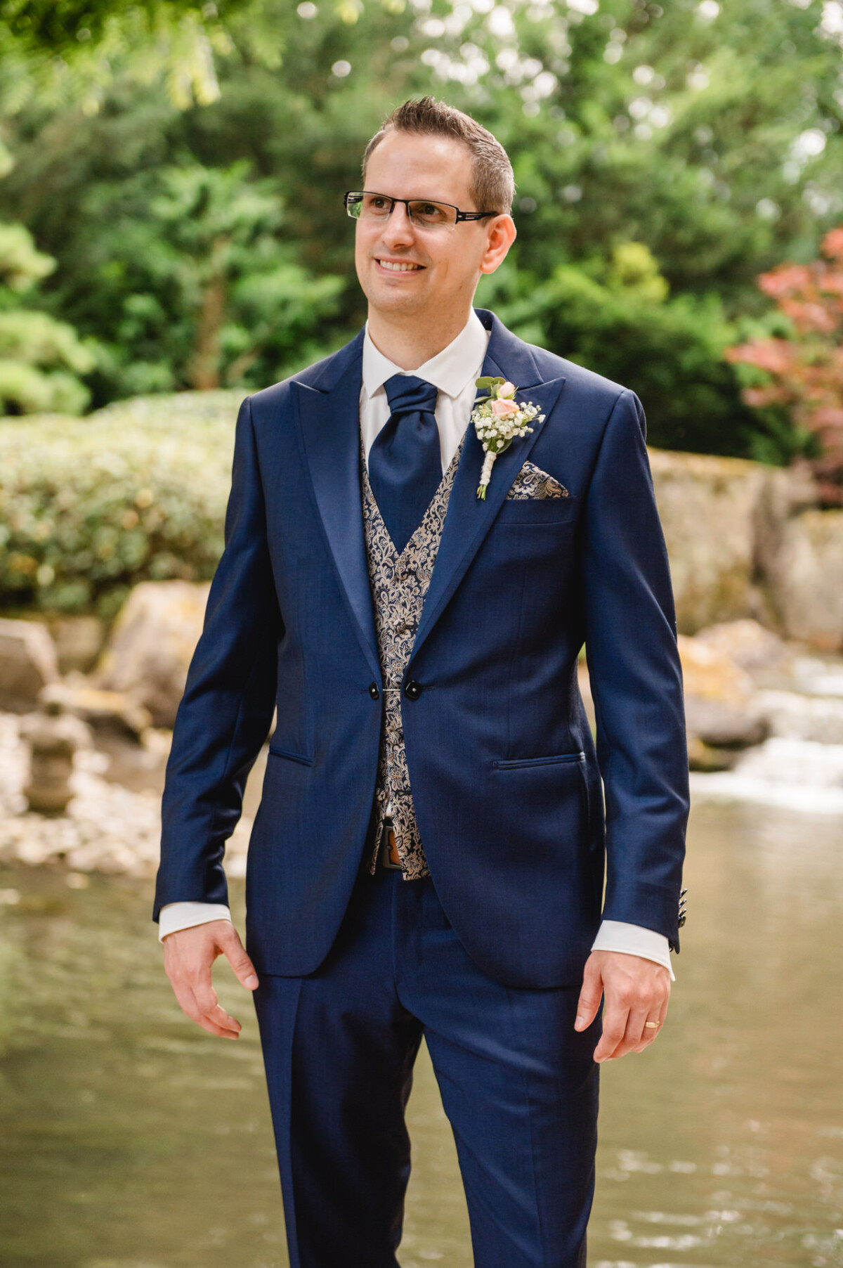 Ein Mann in einem blauen Anzug mit gemusterter Weste steht im Freien neben einem Bach, umgeben von Grün.