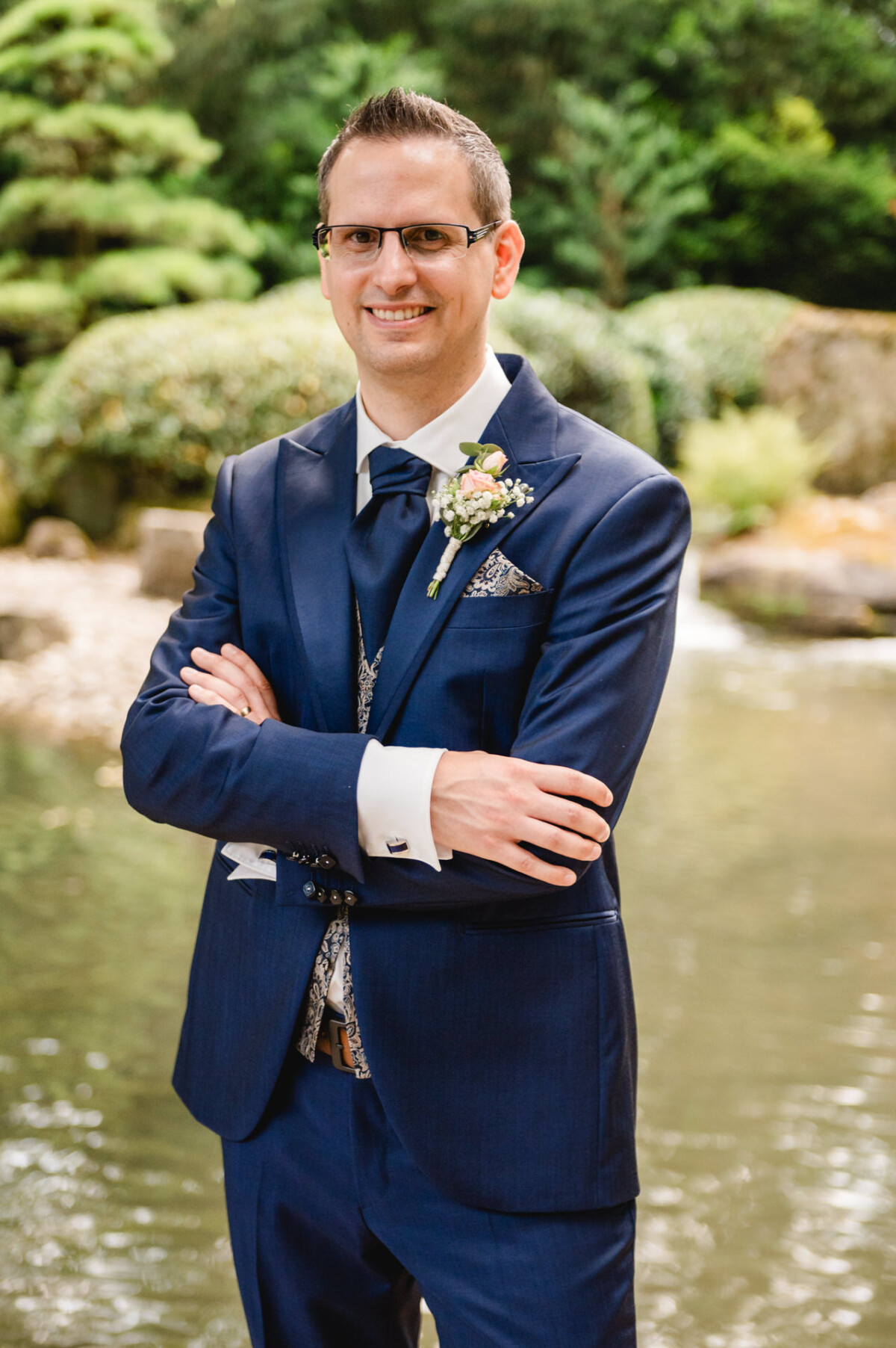 Ein Mann in blauem Anzug und Krawatte steht mit verschränkten Armen vor einem Teich mit Grün im Hintergrund.