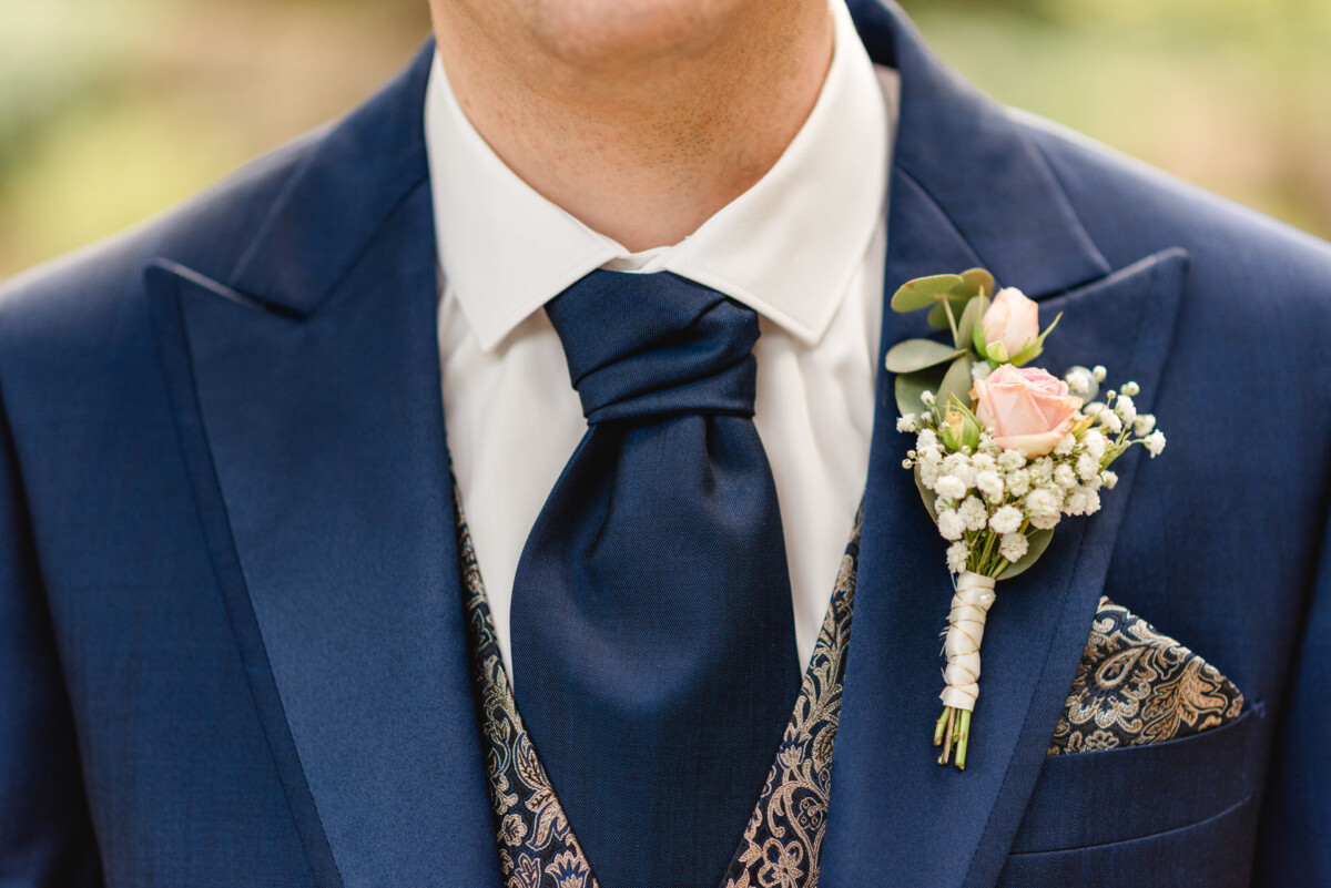 Eine Person trägt einen dunkelblauen Anzug mit einer Weste mit Paisleymuster, ein weißes Hemd und eine blaue Krawatte sowie eine Ansteckblume aus rosa Rosen und Schleierkraut am Revers.