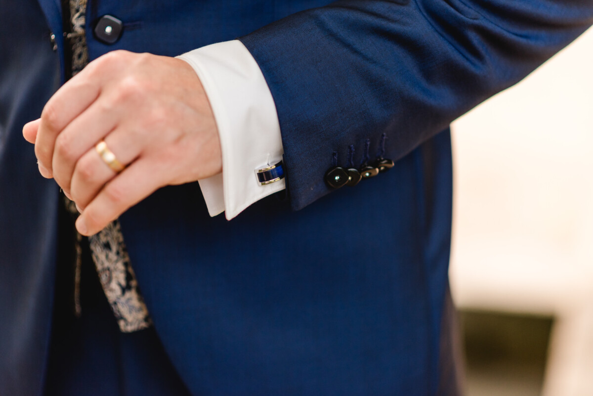 Nahaufnahme einer Person, die den Ärmel ihres Anzugs zurechtrückt. Zu sehen sind eine weiße Manschette mit Manschettenknöpfen und eine Hand mit einem goldenen Ring. Der Anzug ist dunkelblau mit schwarzen Knöpfen.
