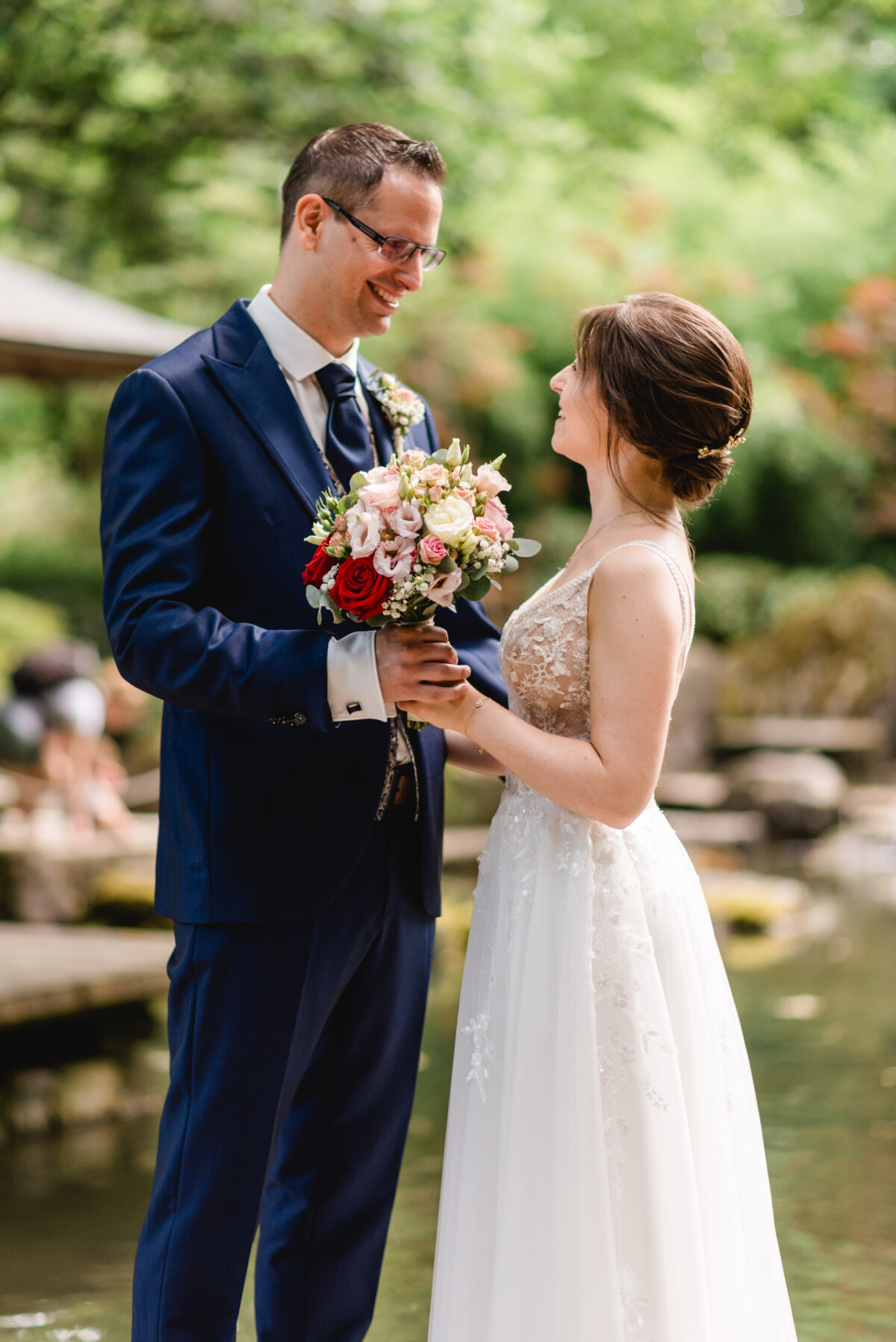 Eine Braut und ein Bräutigam stehen zusammen im Freien, halten Händchen und lächeln einander an, im Hintergrund ist Grün zu sehen.