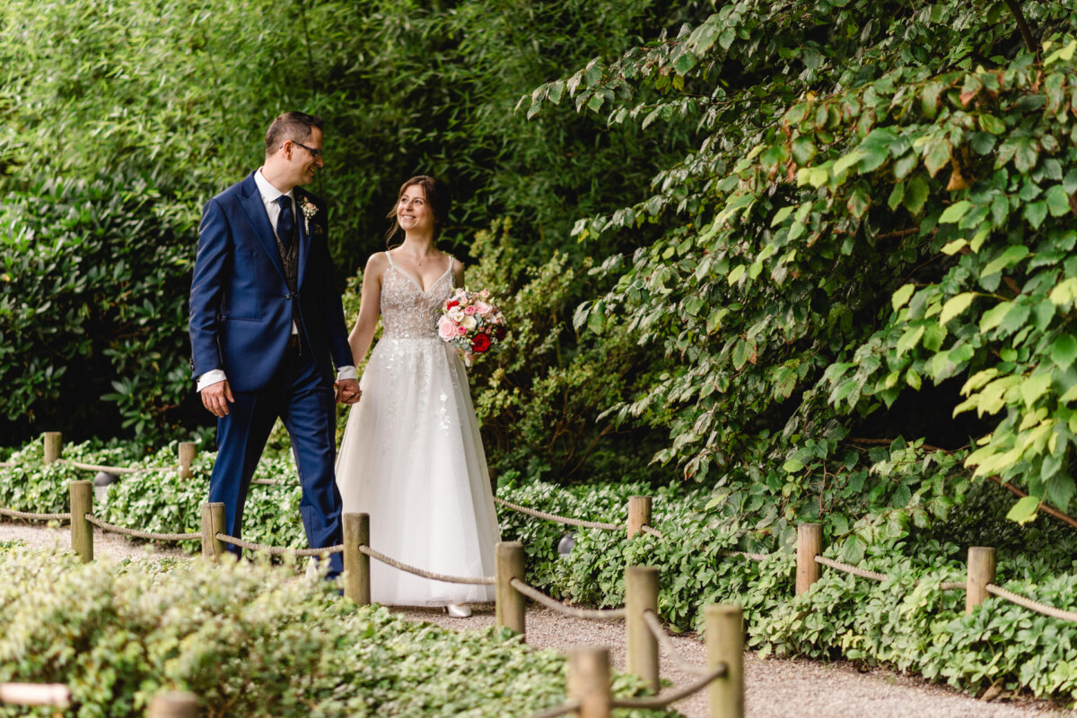 Braut und Bräutigam gehen Hand in Hand einen Gartenweg entlang. Sie hält einen Strauß rosa Blumen in der Hand. Sie sind von üppigem Grün umgeben.
