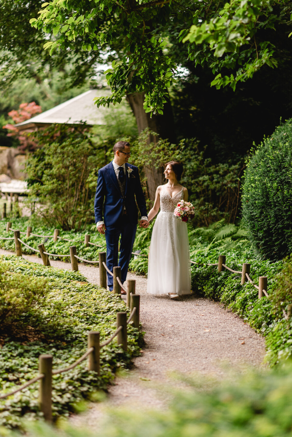 Ein Paar im Hochzeitskleid geht Hand in Hand durch einen üppigen Gartenweg, umgeben von Grün und einem Holzzaun. Die Braut hält einen Blumenstrauß.