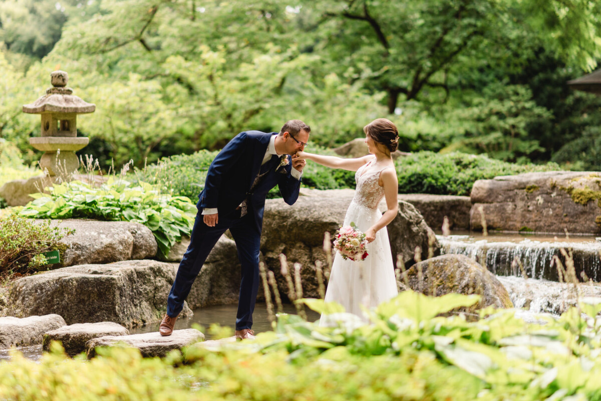 Ein Paar posiert bei einer Gartenhochzeit, wobei der Bräutigam der Braut die Hand küsst. Die Kulisse umfasst Felsen, Grün und einen kleinen Wasserfall im Hintergrund.