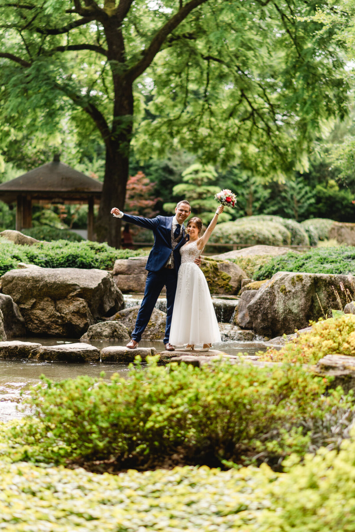 Ein frischvermähltes Paar posiert glücklich auf Felsen neben einem kleinen Wasserfall in einem üppig grünen Park. Die Braut hält einen Blumenstrauß hoch und beide wirken glücklich.