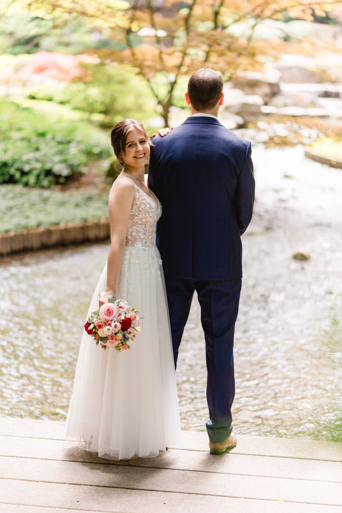 Eine Braut im Spitzenkleid hält einen Blumenstrauß und lächelt in die Kamera, während sie neben einem Bräutigam im blauen Anzug an einem Bach in einem Garten steht.