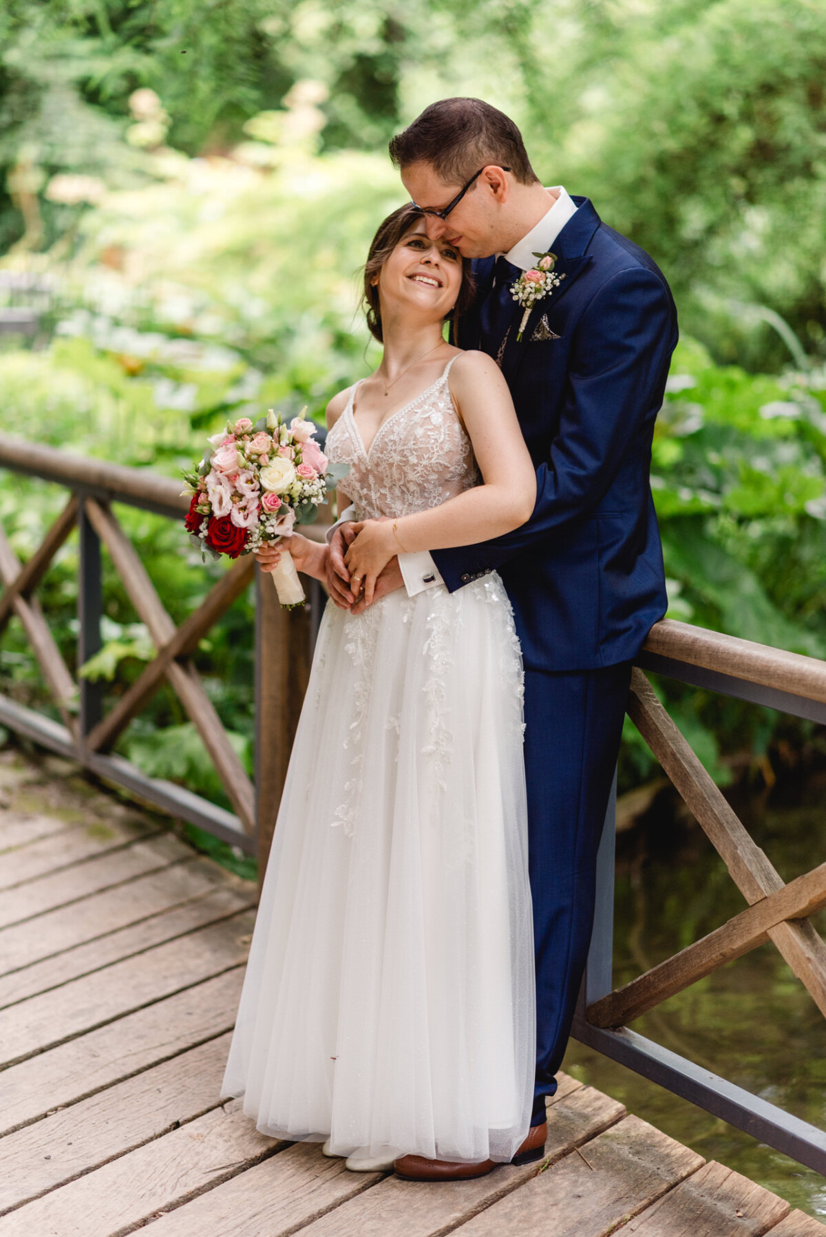 Braut und Bräutigam posieren auf einer Holzbrücke. Die Braut hält einen Blumenstrauß und trägt ein weißes Kleid, während der Bräutigam in einem blauen Anzug sie umarmt. Üppiges Grün umgibt sie.