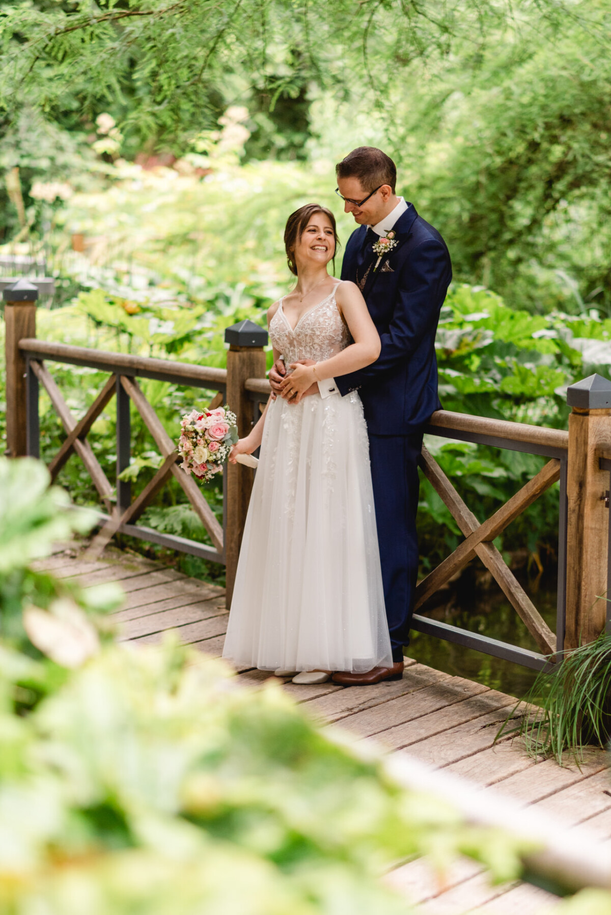 Braut und Bräutigam stehen auf einer Holzbrücke in einem Garten. Die Braut hält einen Blumenstrauß und beide lächeln, während sie einander ansehen.