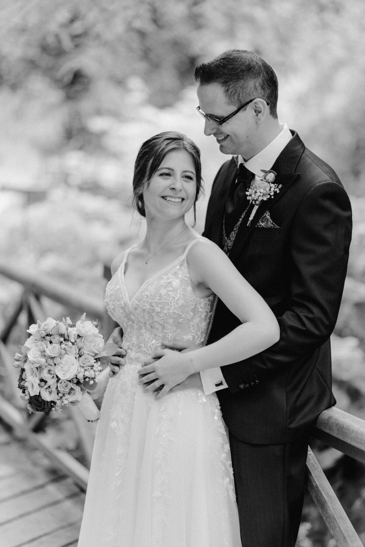 Ein lächelndes Paar steht an seinem Hochzeitstag auf einer Holzbrücke. Die Braut hält einen Blumenstrauß in der Hand und trägt ein Spitzenkleid, während der Bräutigam einen Anzug mit einer Boutonnière trägt.