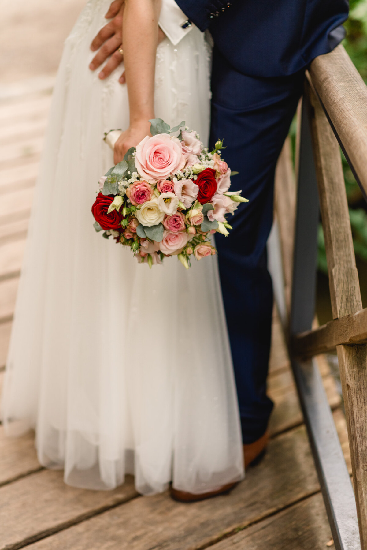 Ein Paar steht eng beieinander auf einer Holzbrücke. Die Frau hält einen Strauß roter, rosa und weißer Rosen. Die Szene fängt den unteren Teil ihrer Körper und das Blumenarrangement ein.