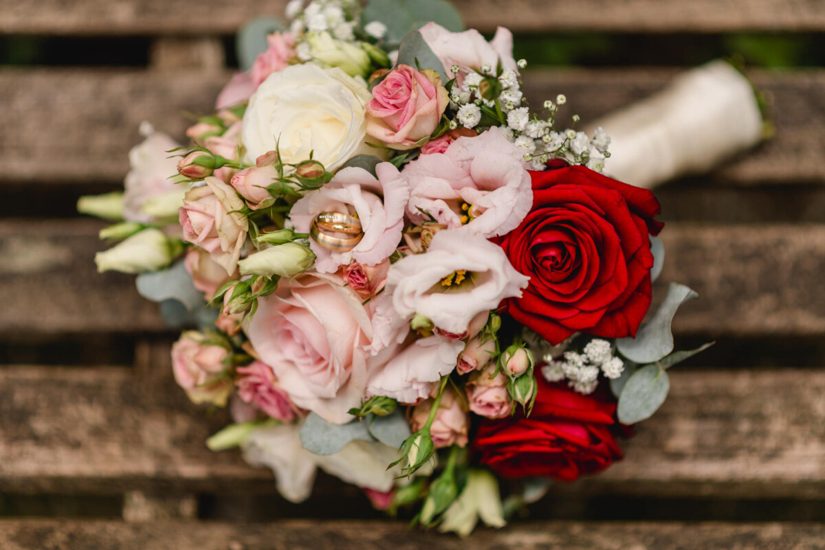 Ein Strauß mit roten, rosa und weißen Rosen mit Grün und kleinen weißen Blüten, auf einer Holzbank platziert.