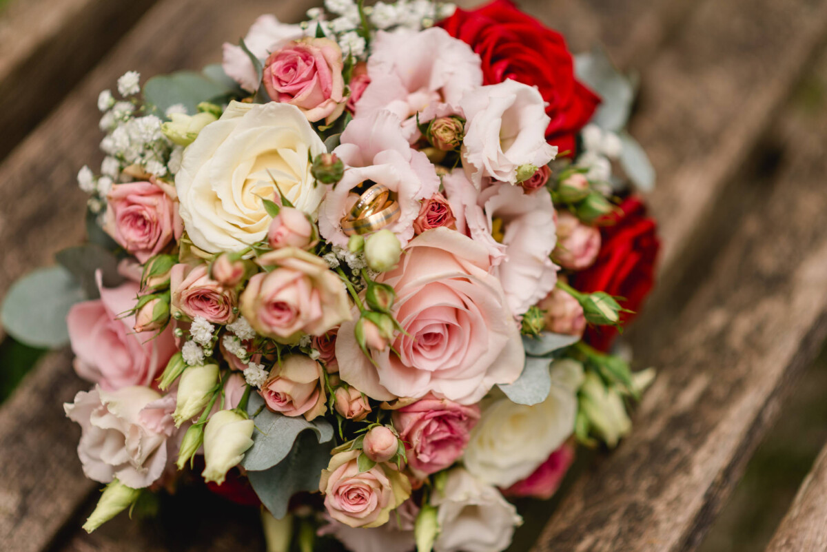 Ein Strauß rosa, roter und weißer Rosen mit Grün auf einer Holzbank.