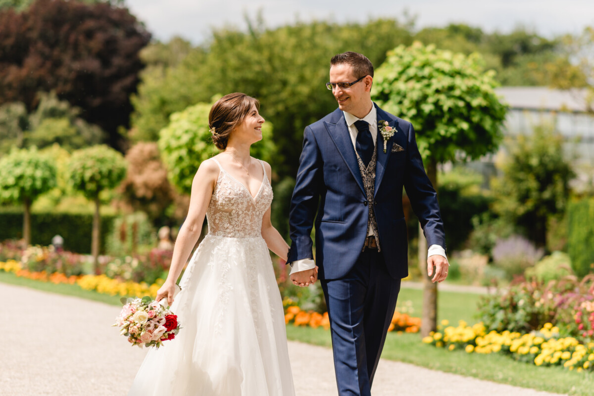 Eine Braut und ein Bräutigam halten Händchen und lächeln sich an, während sie durch einen Garten spazieren.