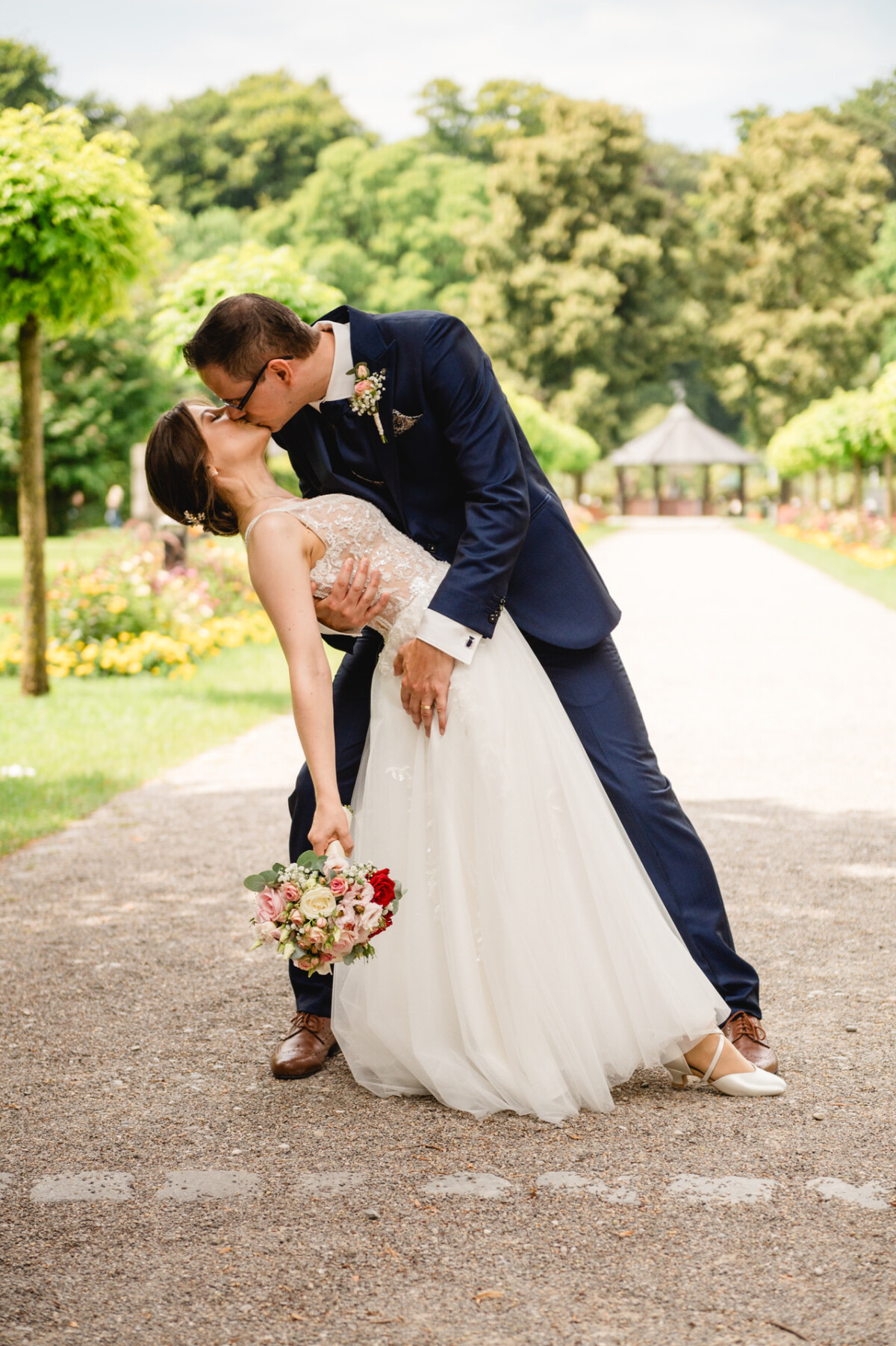 Braut und Bräutigam küssen sich in einem Garten. Der Bräutigam trägt einen blauen Anzug und die Braut hält einen Blumenstrauß, während sie nach hinten geneigt ist.