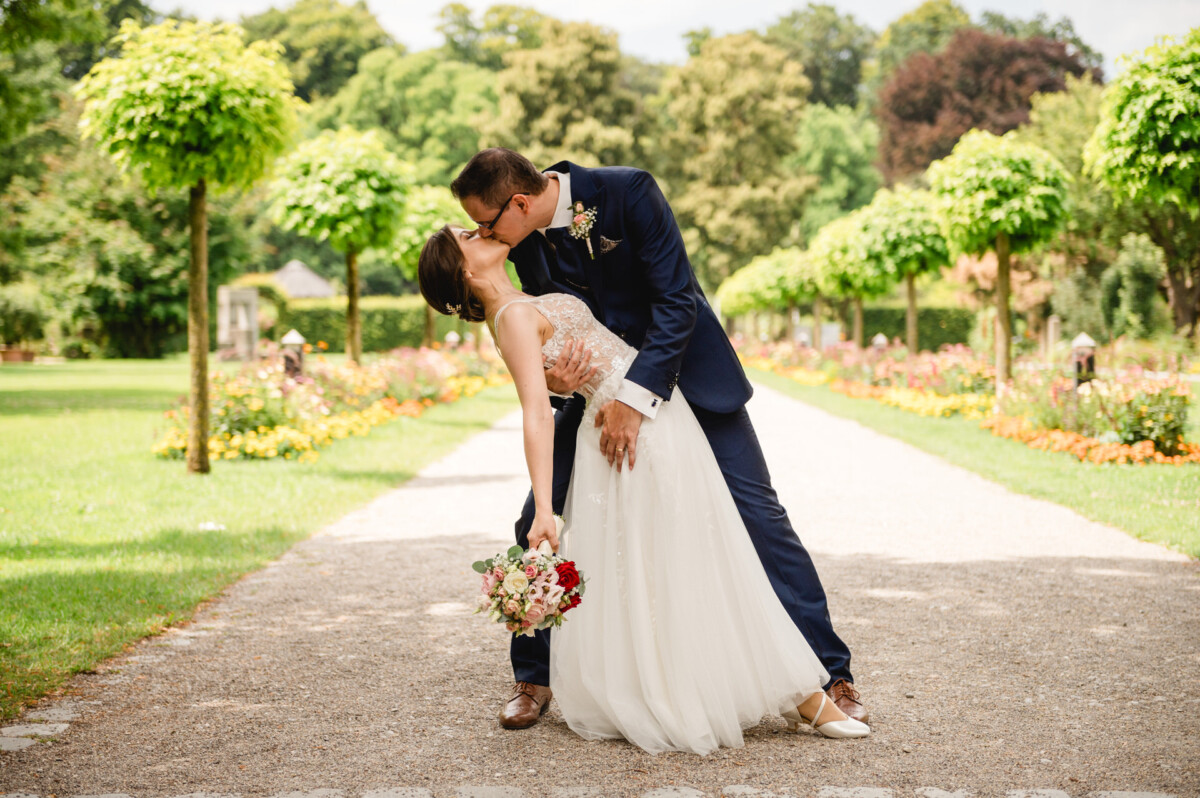 Ein Paar in Abendgarderobe küsst sich auf einem von Bäumen gesäumten Weg. Die Braut hält einen Blumenstrauß und der Bräutigam senkt ihn leicht in die Luft.