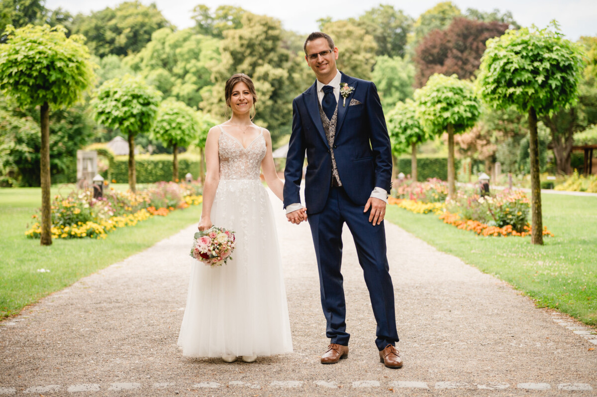 Braut und Bräutigam stehen Hand in Hand auf einem Kiesweg in einem Garten, umgeben von Grün und Blumen.
