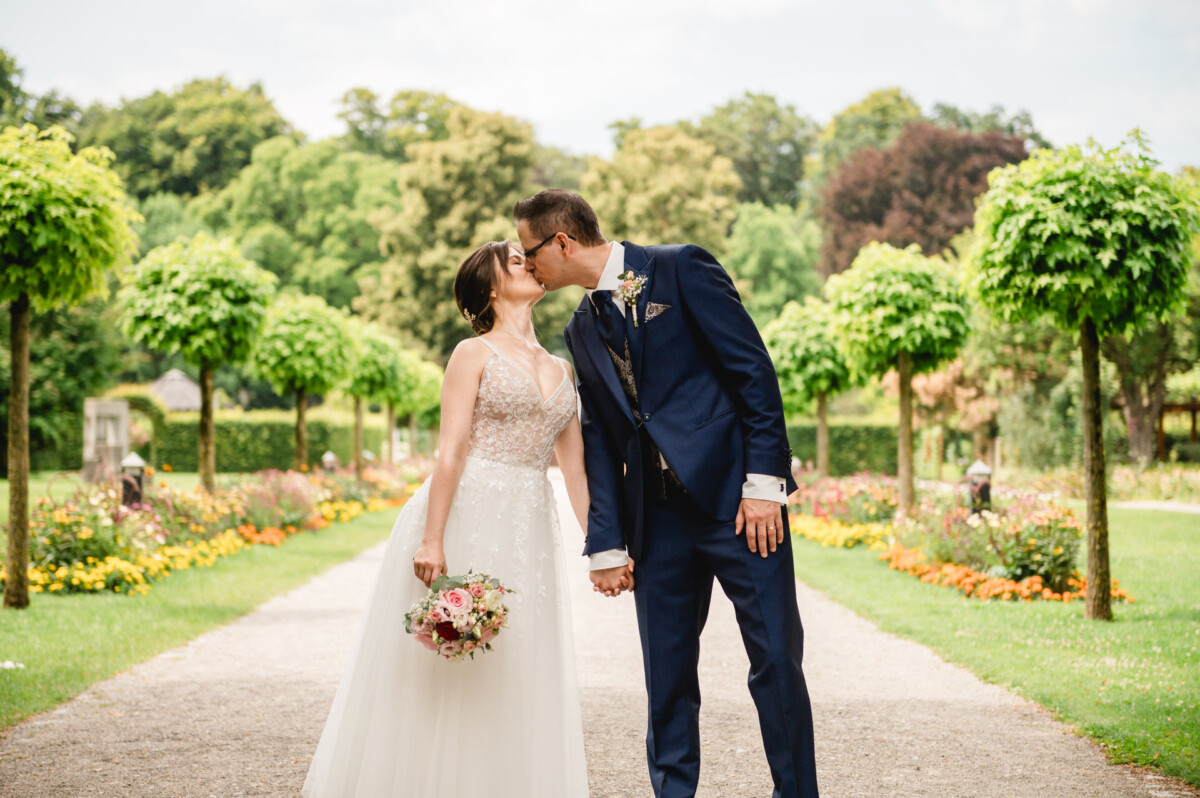 Braut und Bräutigam küssen sich Händchen haltend auf einem von bunten Blumen und Bäumen gesäumten Gartenweg.