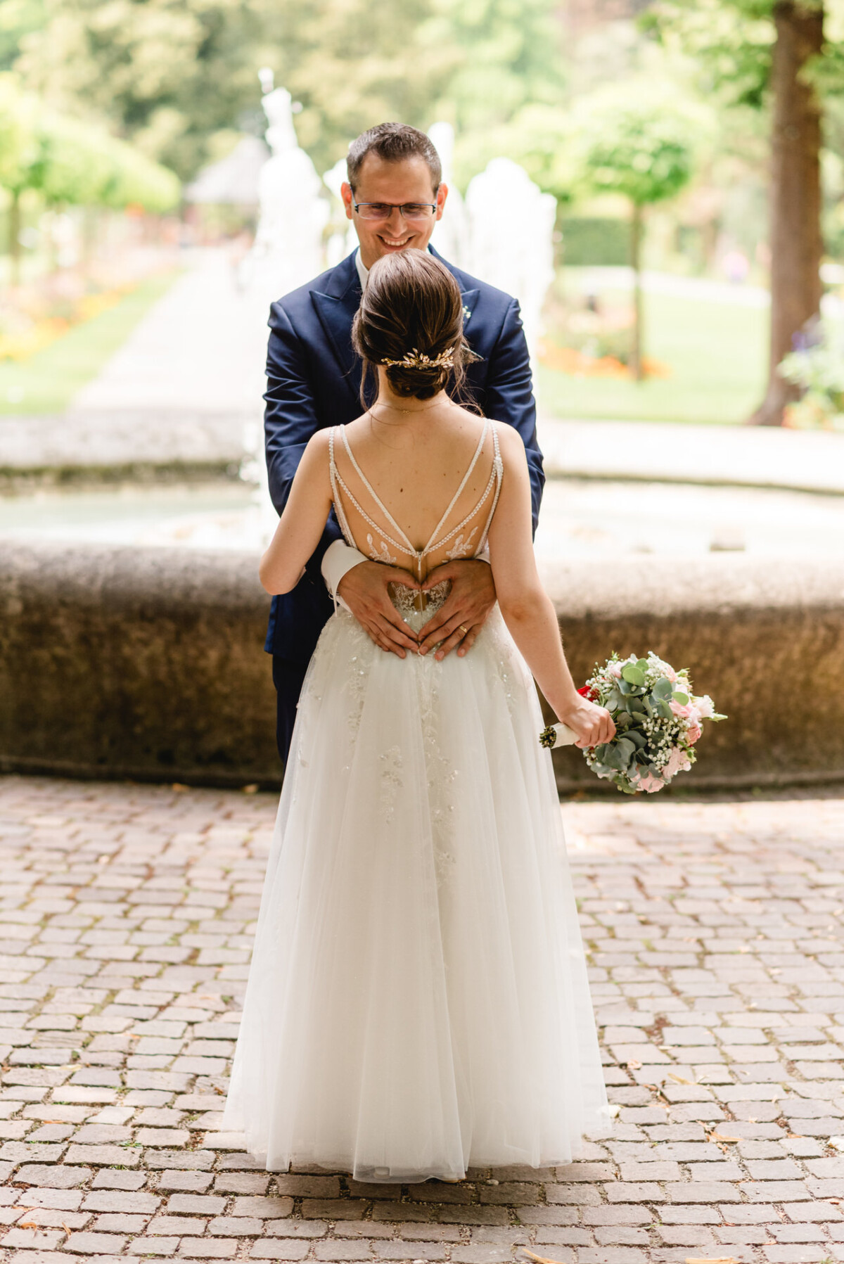 Ein Paar steht eng beieinander in einer Gartenlandschaft neben einem Brunnen. Die Frau im weißen Kleid hält einen Blumenstrauß, während der Mann im Anzug sie umarmt.