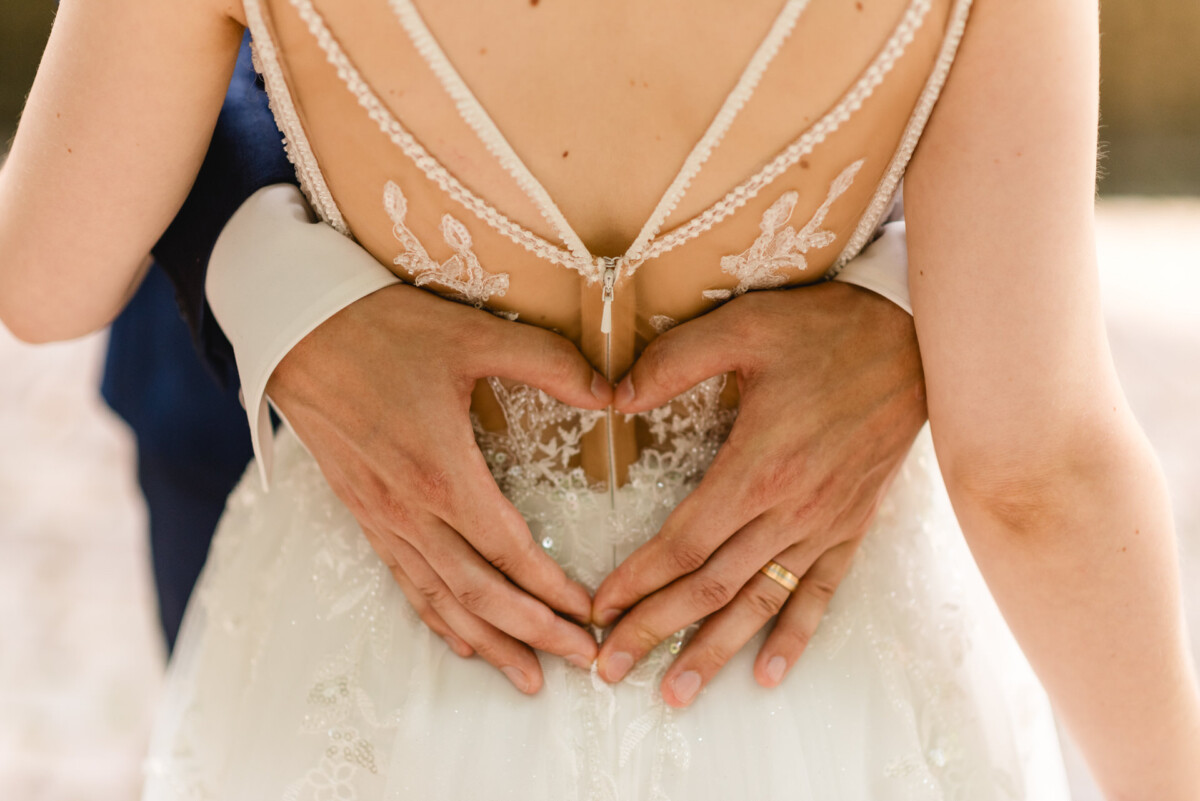Ein Paar formt mit seinen Händen auf der Rückseite des Spitzenbrautkleides der Braut ein Herz.