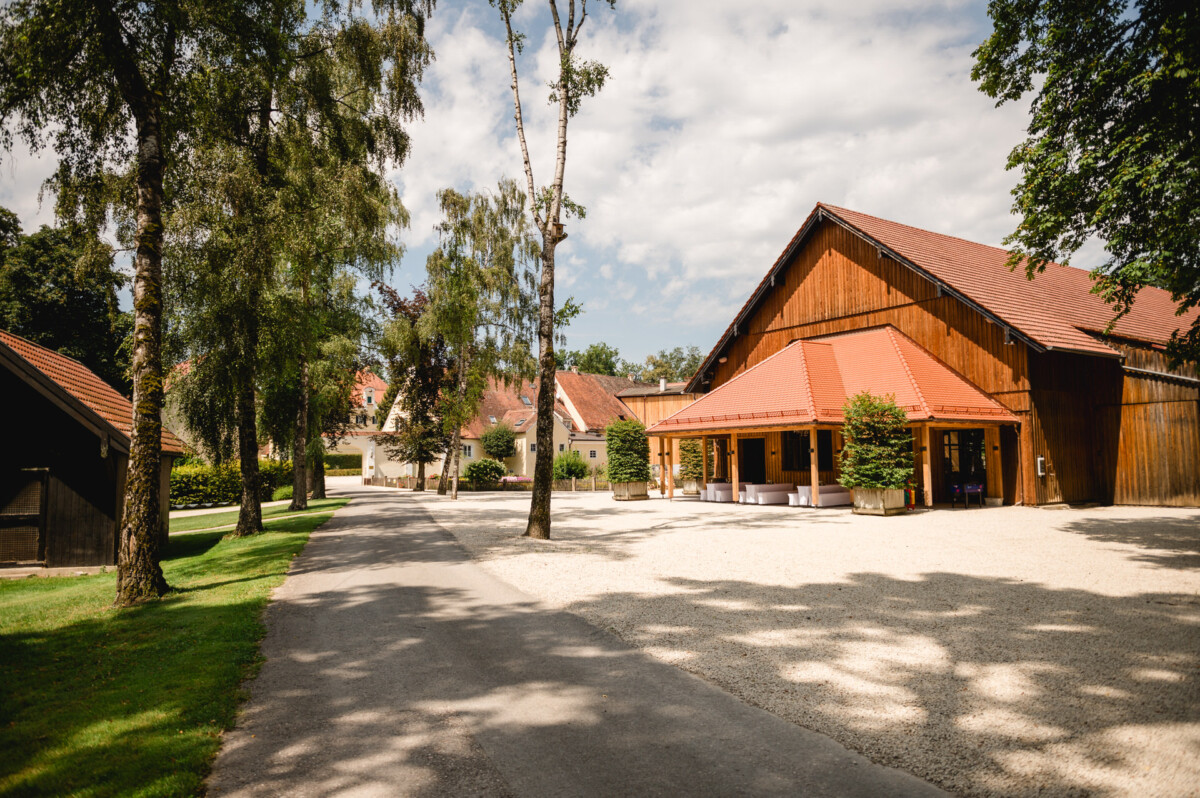 Eine sonnige ländliche Szene mit Holzgebäuden mit roten Dächern und einem von Bäumen umgebenen Kiesweg.