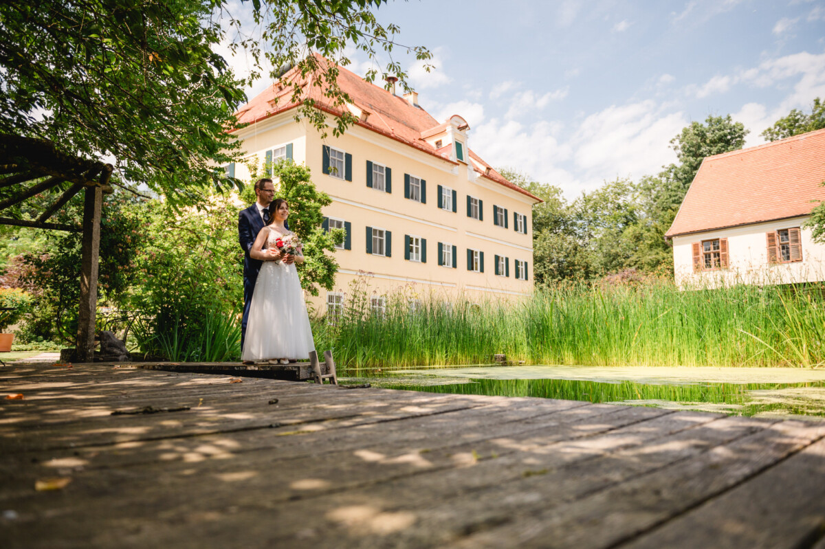 Ein Paar im Hochzeitskleid steht auf einem Holzsteg neben einem Teich, im Hintergrund ist ein gelbes Gebäude zu sehen, und sie sind von viel Grün umgeben.