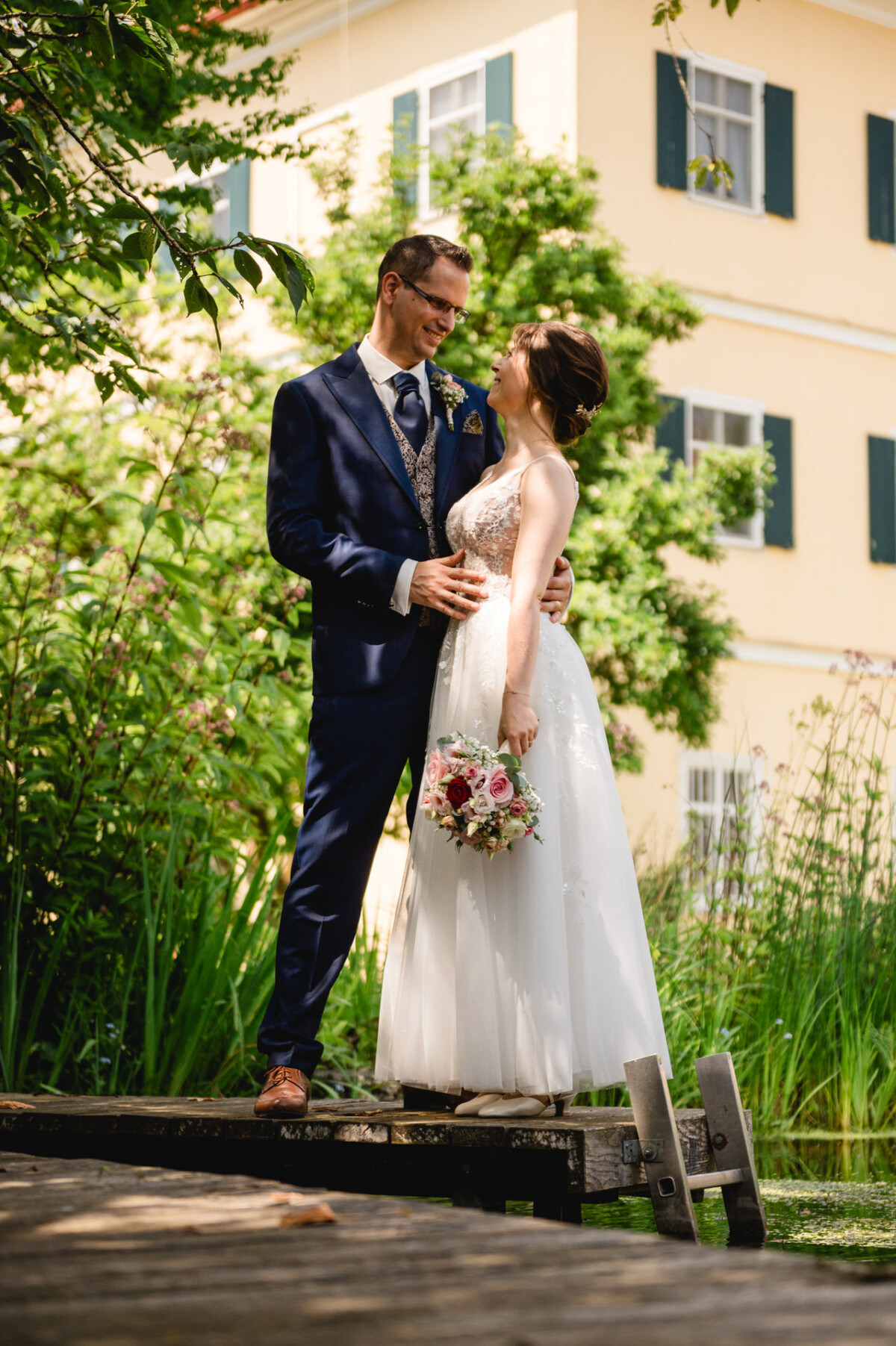 Ein Paar im Hochzeitskleid steht auf einem Holzsteg in einer Gartenlandschaft. Der Bräutigam trägt einen blauen Anzug und die Braut hält einen Blumenstrauß in der Hand. Im Hintergrund ist ein gelbes Gebäude zu sehen.