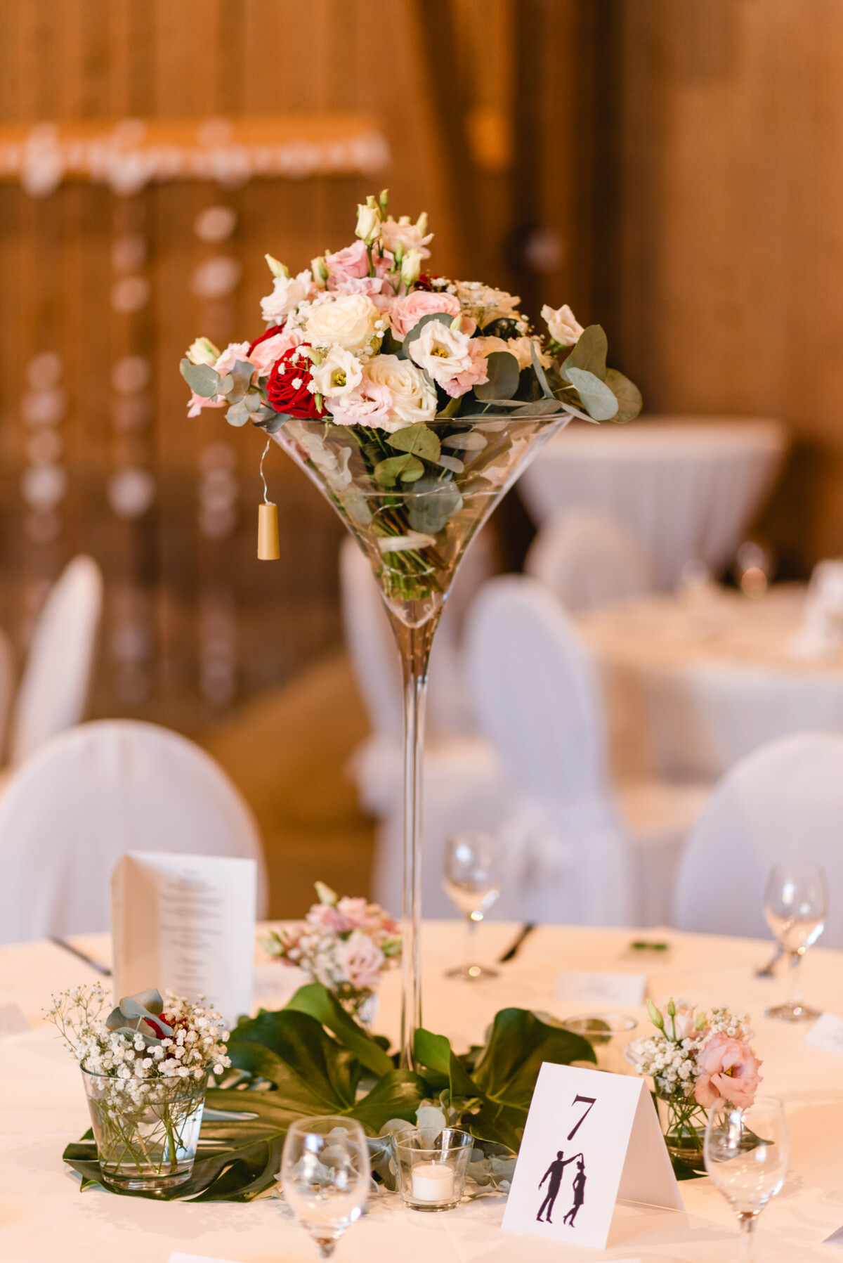 Hoher Blumenschmuck mit rosa und weißen Blumen auf einem runden Tisch, umgeben von kleineren Blumenarrangements und einer Tischnummernkarte mit Silhouetten, im Rahmen einer Hochzeitsfeier.