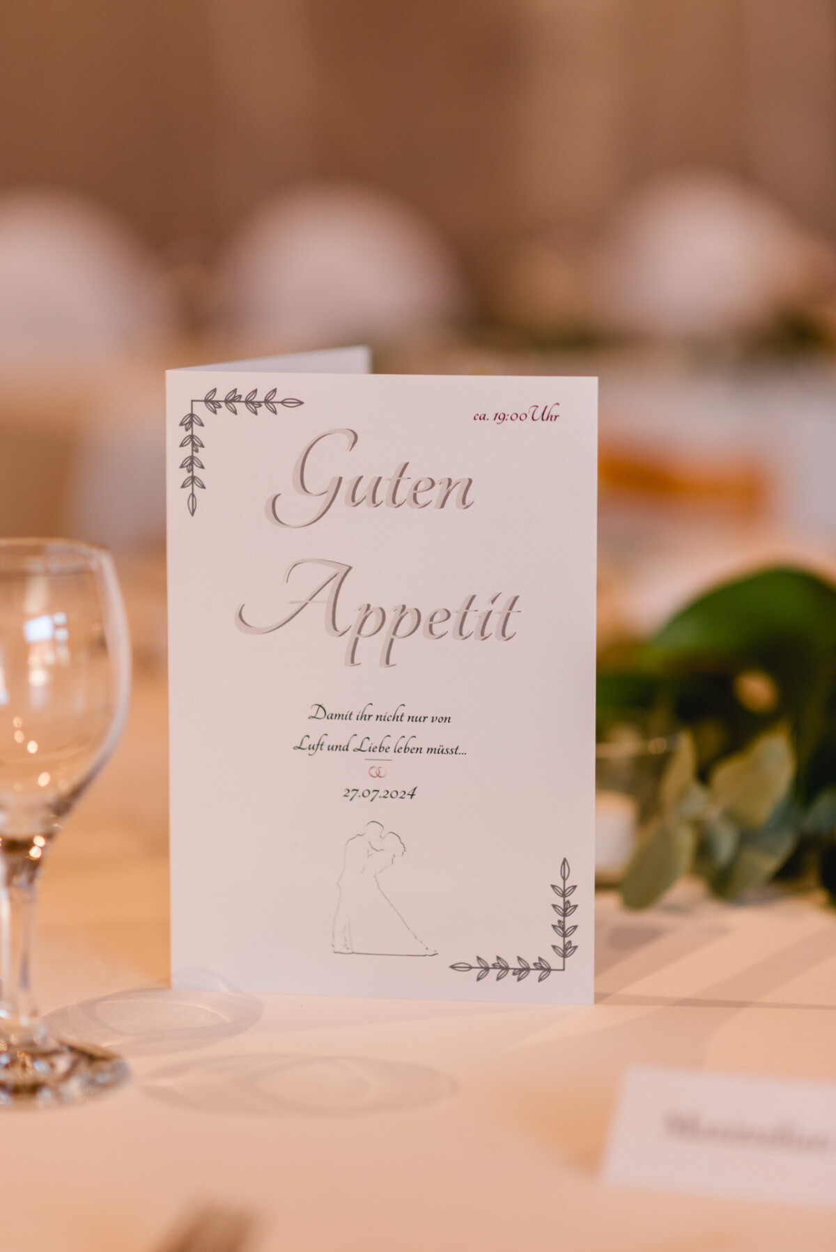 Eine Abendmenükarte mit dem Titel „Guten Appetit“ liegt mit einem Weinglas auf einem Tisch vor unscharfem Hintergrund.