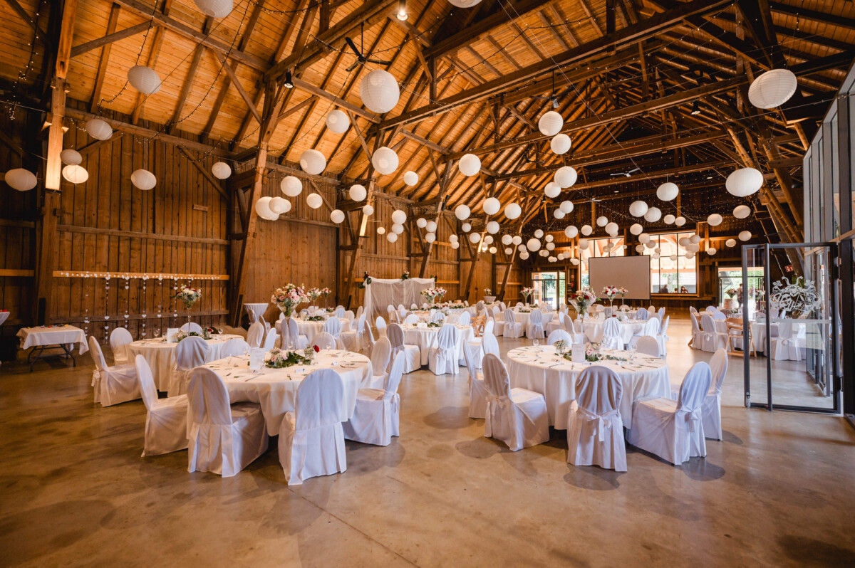 Ein geräumiger Hochzeitsort in einer Scheune mit runden Tischen und weißen Stühlen, dekoriert mit weißen Papierlaternen und floralen Tischdekorationen.